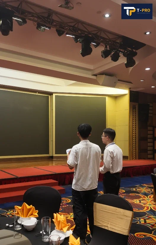 Hai người đứng trước s&acirc;n khấu, ph&iacute;a trước l&agrave; b&agrave;n tiệc v&agrave; ghế trong ph&ograve;ng sự kiện.