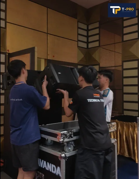 Ba nh&acirc;n sự đang n&acirc;ng một th&ugrave;ng loa cạnh c&aacute;c th&ugrave;ng vận chuyển thiết bị trong ph&ograve;ng sự kiện.